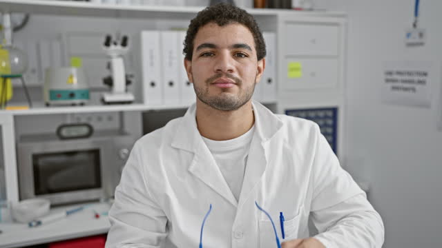 A young adult man with curly hair adjusting safety glasses in a laboratory setting.