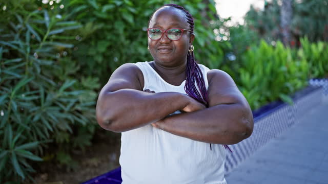 Cool plus-size african american woman, with braids, standing in the calming green park sunlight, arms crossed in serious expression representing a relaxed lifestyle