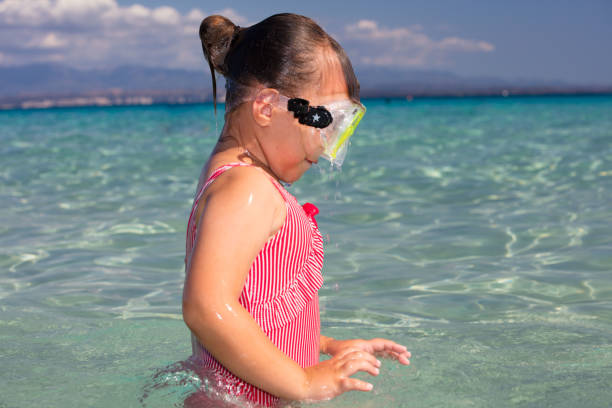 A little girl wearing a diving mask swims in the crystal clear and colorful waters of La Rena Bianca beach in Santa Teresa Gallura.  In the background the Bonifacio Strait and the white cliffs and mountains of Corsica.