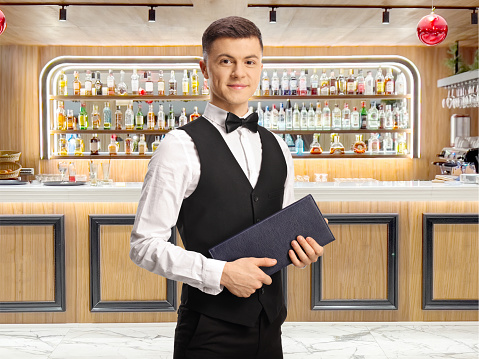 Young server with a bow tie holding a menu in front of a bar with bottles