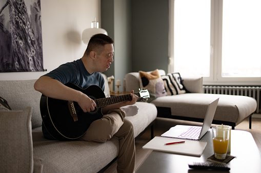 Young man with Down syndrome playing a guitar during an online class over laptop in the living room.