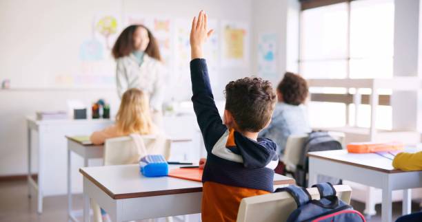 istock Education, raised hand and boy student in classroom with teacher for learning, discussion and knowledge. Help, studying and children students with questions for female educator or tutor at school. 1937157439