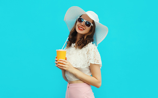 Portrait of beautiful caucasian young woman model with cup of coffee or fresh juice wearing white summer straw hat on studio blue background