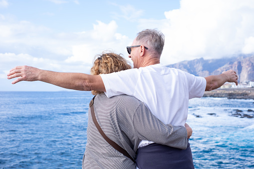 Rear view of mature and romantic senior couple face the sea looking at horizon over water, two smiling people stay together expressing love and tenderness