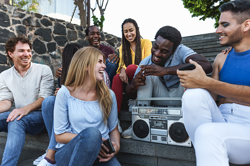 Young multiracial friends having fun listening music with vintage boombox stereo while sitting on city urban stairs - Youth millennial lifestyle concept