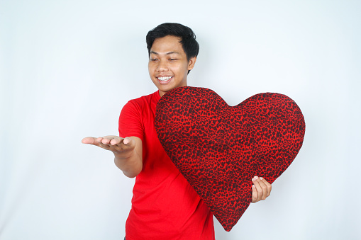 excited asian man holding heart shape pillow presenting with open palm and smiling isolated on white background