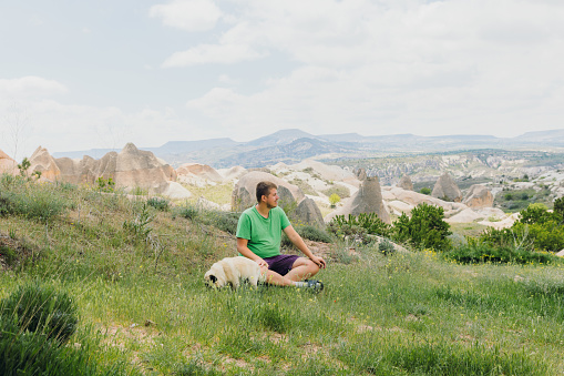 Smiling male explorer enjoying day with his cute pug sitting on the grass surrounded by idyllic rock formations in Nevsehir province, Middle East