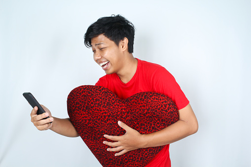 Young Indonesian man holding heart shaped pillow against white background.