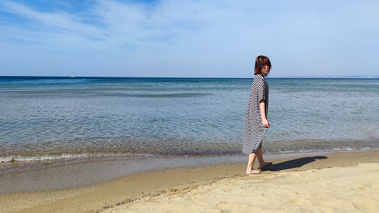 Barefoot teenage girl in a striped dress on the seashore on a sunny day, copy space
