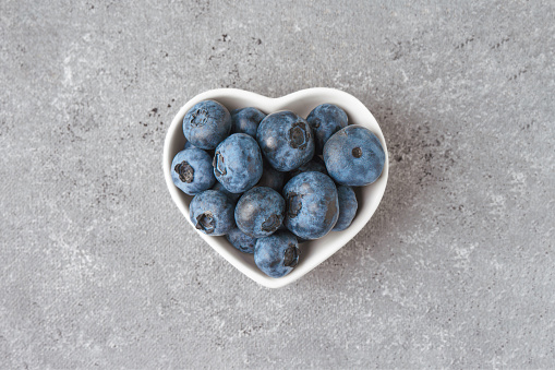 Fresh blueberries in a small heart-shaped bowl on gray concrete background. Organic berries, healthy food, wild berries. Top view, flat lay