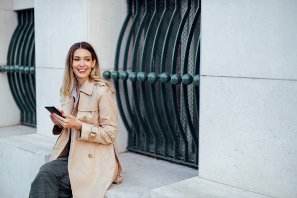 istock A cheerful young lady is sitting on a wall downtown and using her phone. 1937327898
