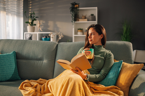 Portrait of a young woman sitting on comfy couch reading a book and drinking coffee or tea in her free time at home. Beautiful female relaxing on a sofa in living room area covered with a blanket.
