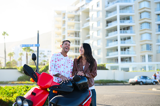 Mature couple riding scooter on tropical beach front with apartment blocks and golden sunlight