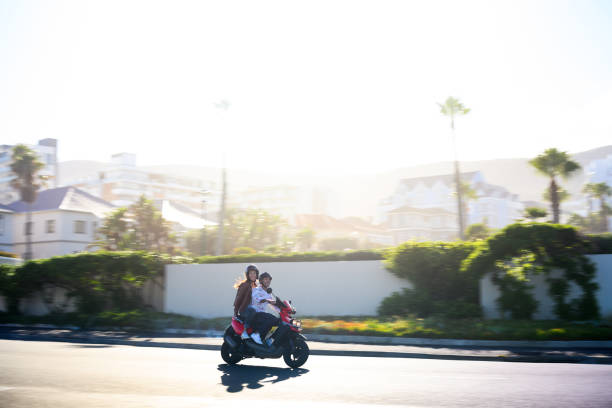 Mature couple riding scooter on tropical beach front with apartment blocks and golden sunlight