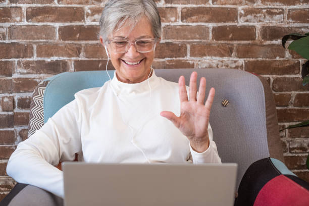 ビデオ通話のコンセプト。ラップトップで手を振る幸せな年配の引退した女性、古い白髪の祖母は、遠くの家族とのオンラインウェブカメラ接続のための技術を楽しんでいます - old armchair women senior adult ストックフォトと画像