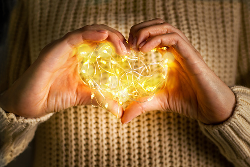 Valentines Day holiday concept. A woman holding hands in shape heart with glowing garland, warm light and bokeh lights. Female in knitted sweater shows a heart-shaped symbol. Love, romance concept.
