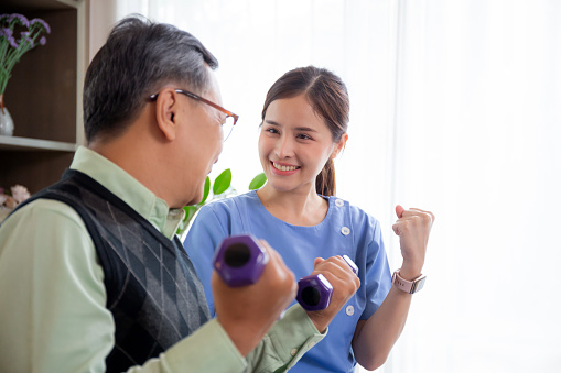 Asian caregiver woman or nurse training senior man lifting dumbbell for arm exercise while therapy and rehabilitation in living room at home, caretaker or physiotherapist helping elderly workout.