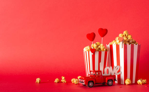 istock Film-inspired Valentine's setup: side view table adorned with miniature car, popcorn boxes, heart decor, sprinkles, against vibrant red wall background. Delightful concept for celebrating love at home 1937429230