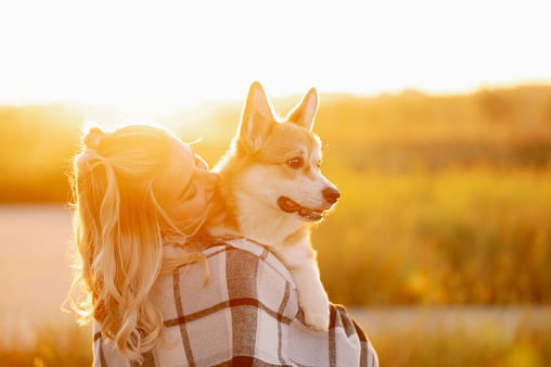 A charming woman sitting in the warm glow of a sunset, wrapping her arms around a joyful corgi dog in a beautiful summer embrace, sharing a moment of pure happiness and affection.