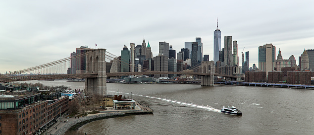 nyc skyline with brooklyn bridge and one world trade center (gray sky, cloudy overcast day) downtown view from manhattan bridge with dumbo carousel and ferry boat on hudson river