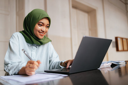 Close up shot of a beautiful Indonesian student wearing hijab, sitting and using a laptop computer. She is at the university doing homework or assignment. She is smiling and looking down at the screen while typing on the keyboard.