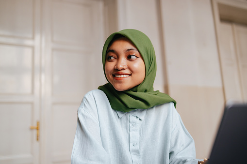 Close up shot of a beautiful Indonesian young woman wearing a green hijab. She is sitting, smiling and looking away.