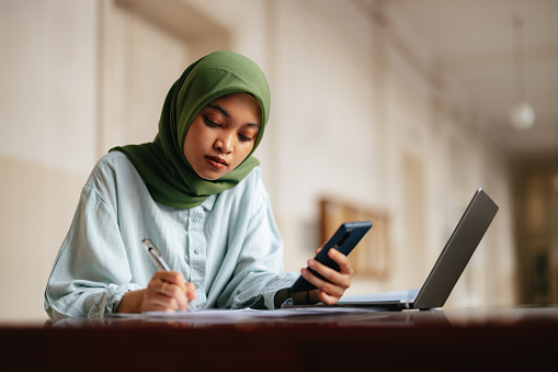 Close up shot of a beautiful serious young woman wearing hijab, sitting at the desk in the school's hallway, studying or doing homework while holding mobile phone. She is looking down at the paper and writing.