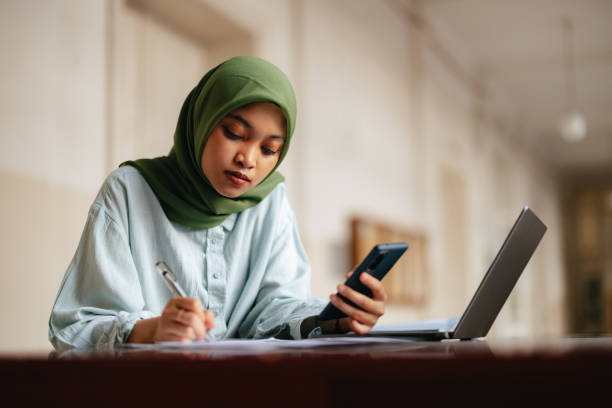 portrait of a beautiful serious indonesian female student holding mobile phone while doing homework - homework teenager mobile phone school imagens e fotografias de stock