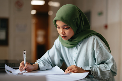 Portrait of a beautiful young serious woman wearing hijab, sitting and writing and assignment for a school project or homework. There is a laptop computer on the desk.