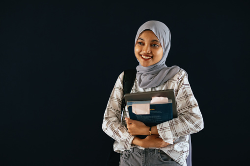 Portrait of a beautiful Indonesian female student wearing grey hijab, standing by the black wall, holding a laptop computer and a textbook, smiling and looking away.