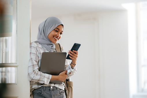 Portrait of a beautiful young smiling Indonesian woman wearing hijab, standing in the school's hallway leaning against the wall. She is holding and using a mobile phone for texting or reading a message.