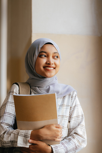 Portrait of a beautiful young Indonesian woman wearing hijab, holding books in her arms, standing and leaning against the wall in school. She is smiling and looking away.