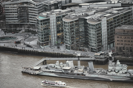 London, England, United Kingdom, Great Britain – January 2024: More London Riverside Buildings (Business centre) and the HMS Belfast (museum ship, a Town-class light cruiser that was built for the Royal Navy, the United Kingdom's naval warfare force) on the river. An aerial photograph showing the urban panorama view/city skyline taken in the heart of the financial district of the City of London. This photo was captured at London's East End.