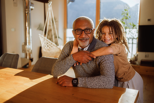 In a room filled with the soft light of the setting sun, a grandfather with glasses shares a warm hug with his young grandson. Both are smiling, enjoying a simple, happy moment together, surrounded by the comfort of their home.