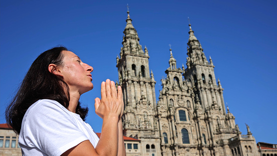 a poignant moment of a woman in devout prayer, her hands pressed together and her eyes closed in reverence, with the majestic Santiago de Compostela Cathedral looming in the sharp blue sky behind her. The image encapsulates a personal journey of faith, pilgrimage, and the emotional culmination at one of Christianity's most sacred sites.