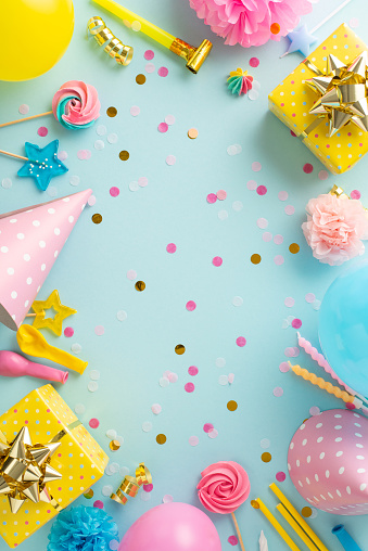 Exciting birthday concept composition. Overhead vertical image of festively decorated table with gifts, birthday hats, balloons, confetti, and more against soft blue backdrop. Ideal for text or advert