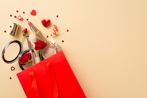 Unwrap romance! Top view of paper bag bursting with gifts: lipstick, brushes, eyeshadow, powder, red roses, hearts and confetti. Against a pastel beige backdrop, ready for your heartfelt message