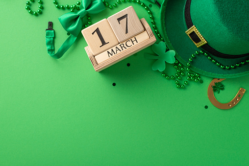 St. Patrick's celebration scene: Top view of the date on a wooden calendar, leprechaun's hat, lucky horseshoe, trefoils, confetti, and beads on a vibrant green background
