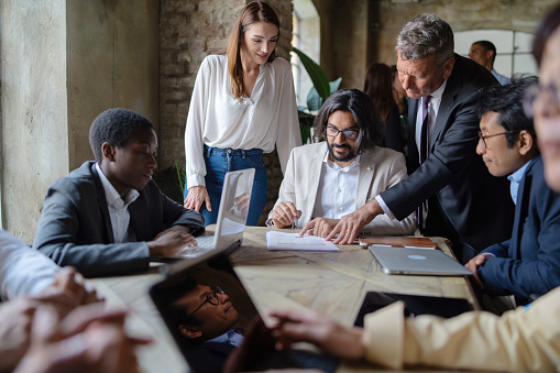 A multicultural business team is gathered around a table, laptops at hand. A young Middle-Eastern entrepreneur signs documents guided by a senior in suit and tie.