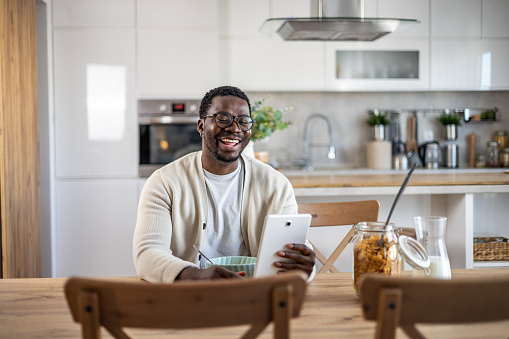 A smiling young black man is looking at something on a tablet while enjoying a healthy breakfast of cornflakes and milk