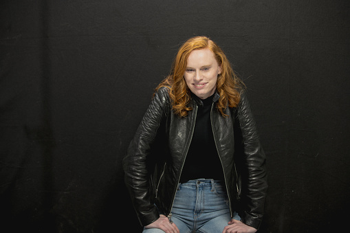 Young Los Angeles citizen with red hair, wearing a black sweater, jeans and a black leather jacket poses in studio before a black background