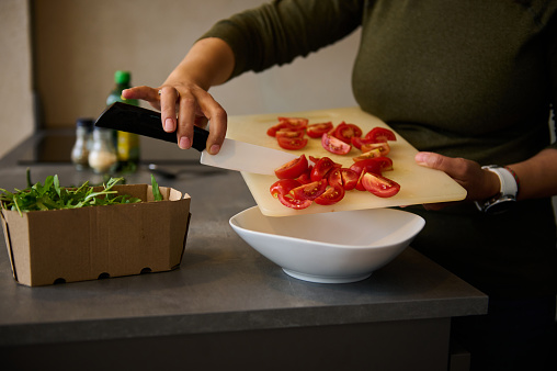 Chef hands with knife, slicing and pouring red tomatoes from cutting board into white bowl for salad, cooking healthy meal for lunch. Fresh vegetables, arugula leaves and olive oil on kitchen table