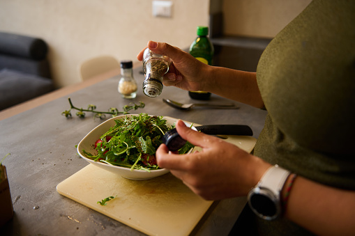 Close-up of a young woman preparing healthy vegetable salad in kitchen, seasoning meal, adding some herbs and salt to bowl, enjoying cooking vegetarian food at home, free space for advertising text