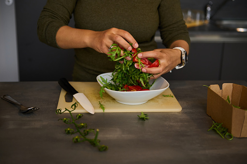 Selective focus on hands of a woman chef, housewife, standing at kitchen counter, holding fresh green arugula leaves and juicy red tomatoes while preparing healthy raw vegan salad in the home kitchen