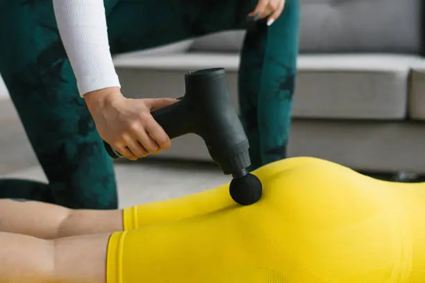 Female massage therapist does massage exercises on the hips with the help of a shock vibrator massager to a female client at home. Shock therapy for a restorative massage of the athletic body