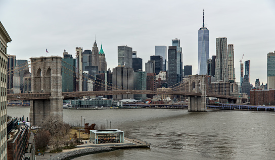 nyc skyline with brooklyn bridge and one world trade center (gray sky, cloudy overcast day) downtown view from manhattan bridge with dumbo carousel and ferry boat on hudson river