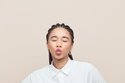 Playful multiracial teenage girl with braided hair wearing white blowing kiss at camera with eyes closed . Studio shot, beige background.