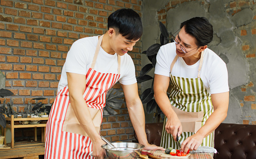 Two asian handsome men wearing apron, casual clothes, cooking together in kitchen room at home, smiling with happiness. Lifestyle, Friendship and LBGT Concept