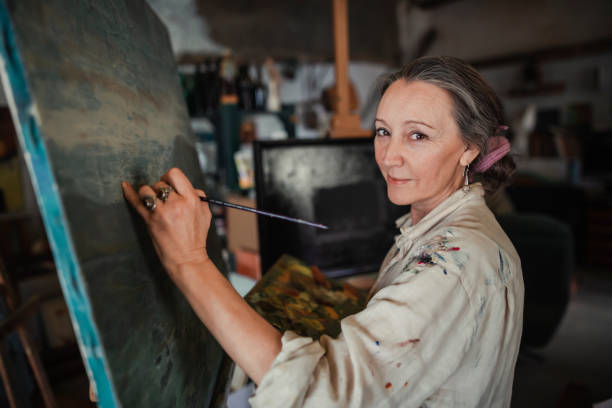 mujer pintando un cuadro mientras mira a la cámara - oficio artístico fotografías e imágenes de stock