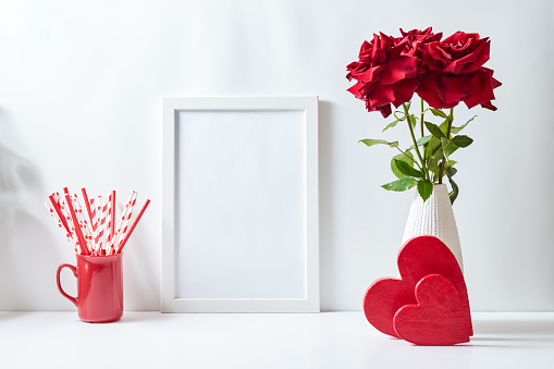 Mockup with a white frame and red roses in a vase, red heart  on a white table. Empty poster frame mockup for presentation, design, lettering